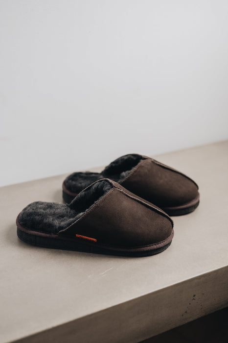 Pair of dark brown sheepskin slippers on a light concrete surface with a neutral background