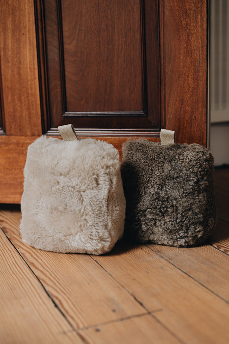 A wheat and oatmeal door stop with leather tabs, holding a wooden door open, on a wooden floor