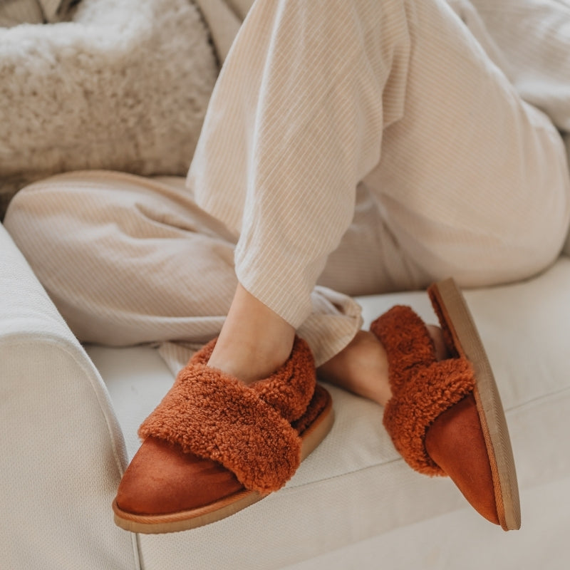 Rust Coloured sheepskin slippers worn with beige pants on a light-colored floor.