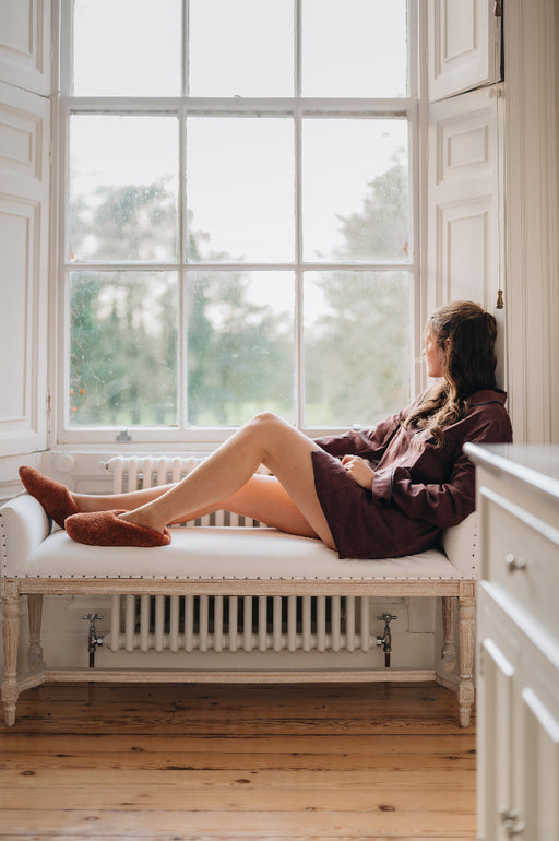 Young woman sat in window seat wearing curly merino sheepskin slip on slipper with curls on the outside and inside in a rich, warm rust colour.