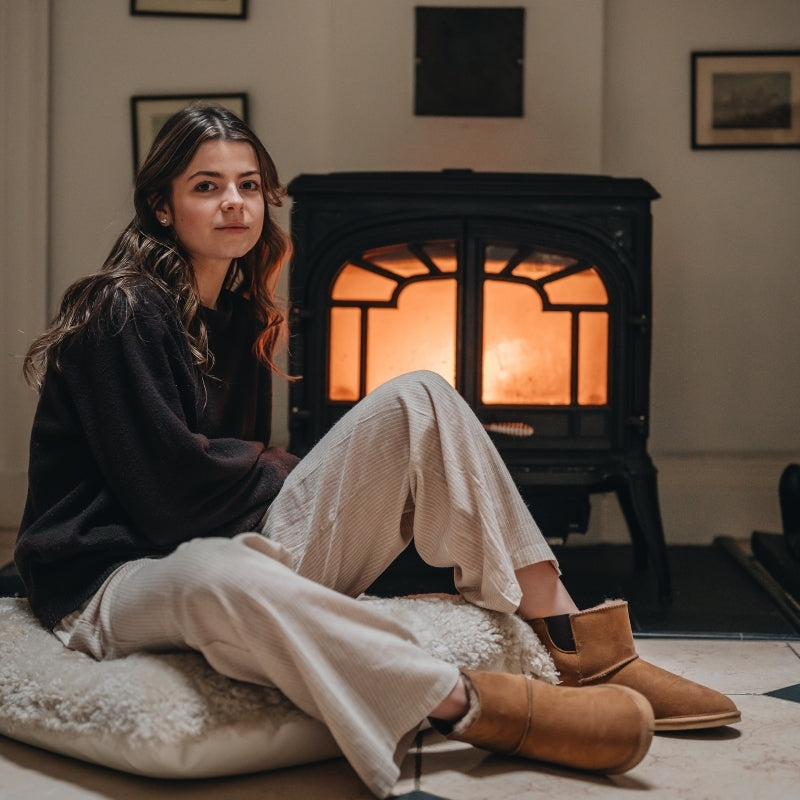 Woman sitting in front of a lit fireplace wearing a dark sweater, light pants, and sheepskin slipper boots.