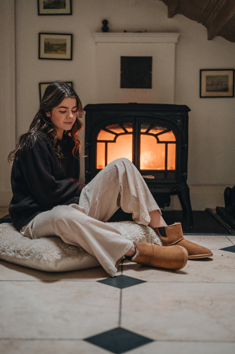Young woman sitting on a sheepskin floor cushion in front of a log fire wearing the Winnie sheepskin slipper boot. She looks peaceful and relaxed.