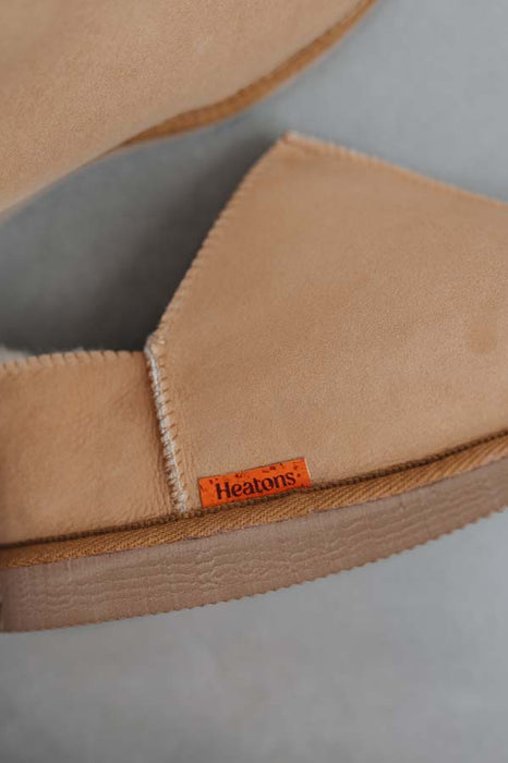 Close-up of a tan pair of Cadi sheepskin slippers with a small orange Heaton's brand label, resting on a neutral grey surface.