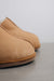 A close-up shot of a pair of tan sheepskin 'Cadi' slippers on a neutral grey surface against a light background.