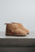 A pair of tan merino sheepskin ankle slipper boots with a small orange cork Heatons brand tag, displayed on a grey concrete bench.