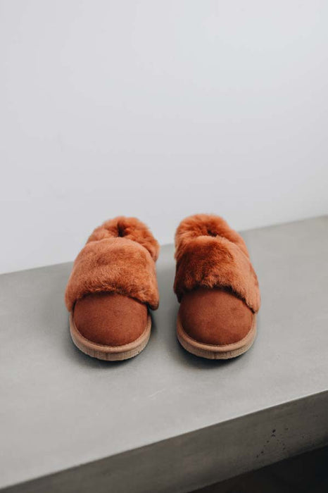 Front view of a pair of rust coloured sheepskin slippers on a light grey surface with a white background