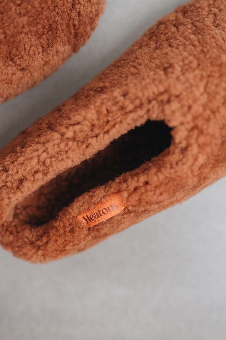 Close up of rust coloured curly sheepskin slippers with 'Heatons' brand logo on an orange cork label