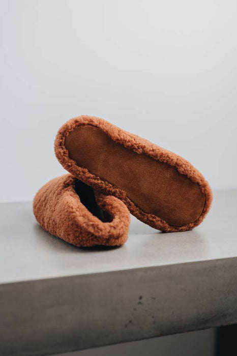 Suede underside of a pair of rust curly sheepskin slippers on a light grey stone bench with a neutral background