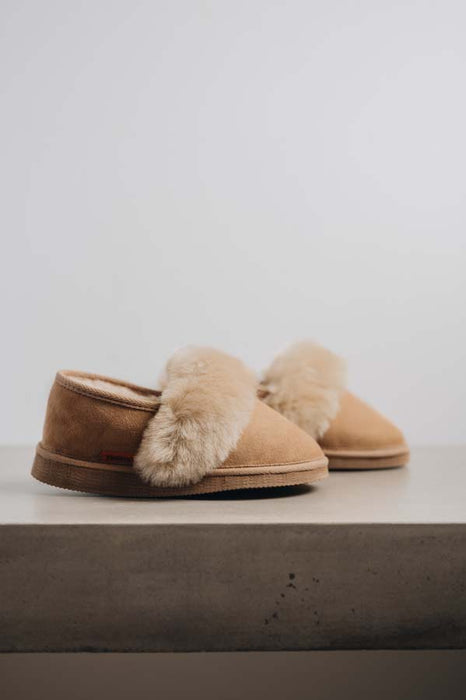 A pair of tan merino sheepskin slippers with fluffy cream shearling trim, displayed on a light grey concrete surface against a neutral light grey background.