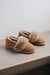 A pair of taupe coloured sheepskin slippers with fluffy shearling cuff, resting on a light grey concrete surface against a neutral off-white background.