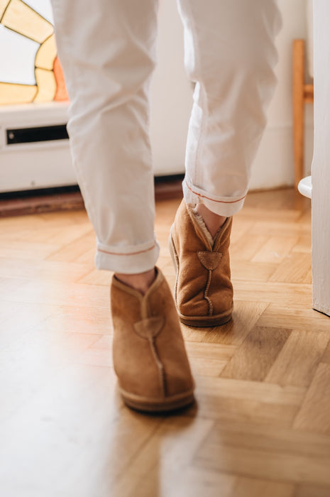 Men's boot slippers with sheepskin with hard sole. Named Huw and rafted from Spanish Merino. Worn by man walking away from a stained glass door on an oak floor.