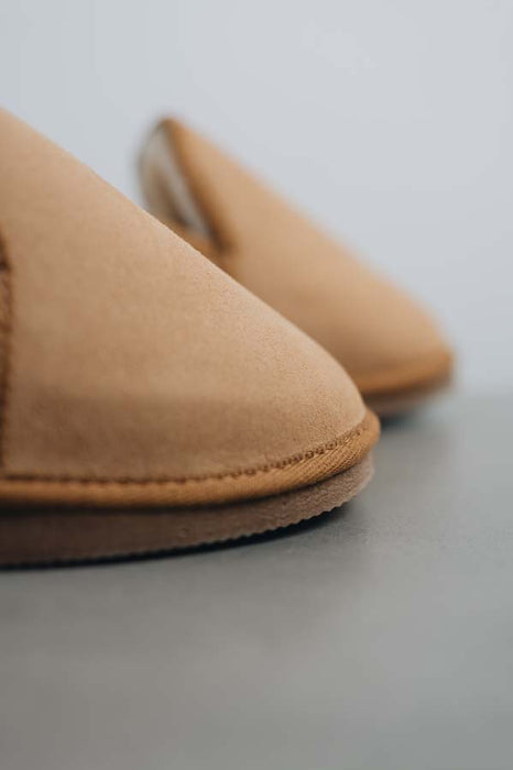 Close-up of a pair of tan sheepskin slippers on a neutral grey surface, with one slipper in sharp focus and the other softly blurred in the background.