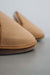 Close-up of a pair of tan sheepskin slippers on a neutral grey surface, with one slipper in sharp focus and the other softly blurred in the background.