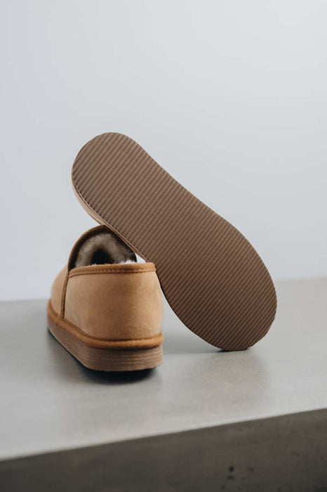 A tan men's sheepskin slipper shown from the back with its sole propped up, revealing a ribbed brown outsole