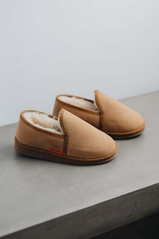 A pair of men's tan sheepskin slippers with a soft cream fleece lining, resting on a grey concrete ledge against a light gray wall.