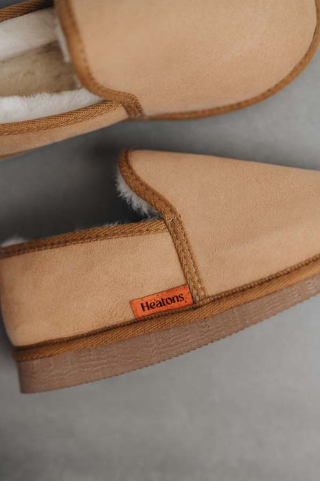 Close-up of a pair of Reggie men's sheepskin slippers with white shearling lining and a small orange Heatons brand tag on the side, set against a neutral grey background.