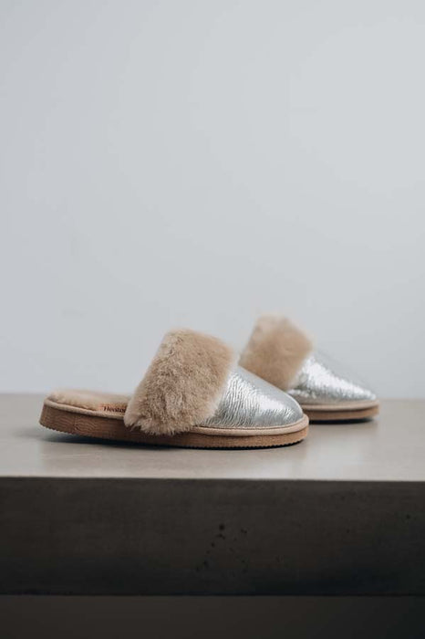 A pair of open-toe slippers with metallic silver uppers and fluffy beige genuine sheepskin trim, resting on a stone surface against a light grey background.